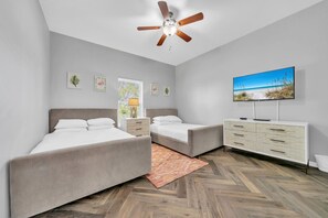 A modern bedroom with two beds, a ceiling fan, a TV, and a herringbone wood floor.