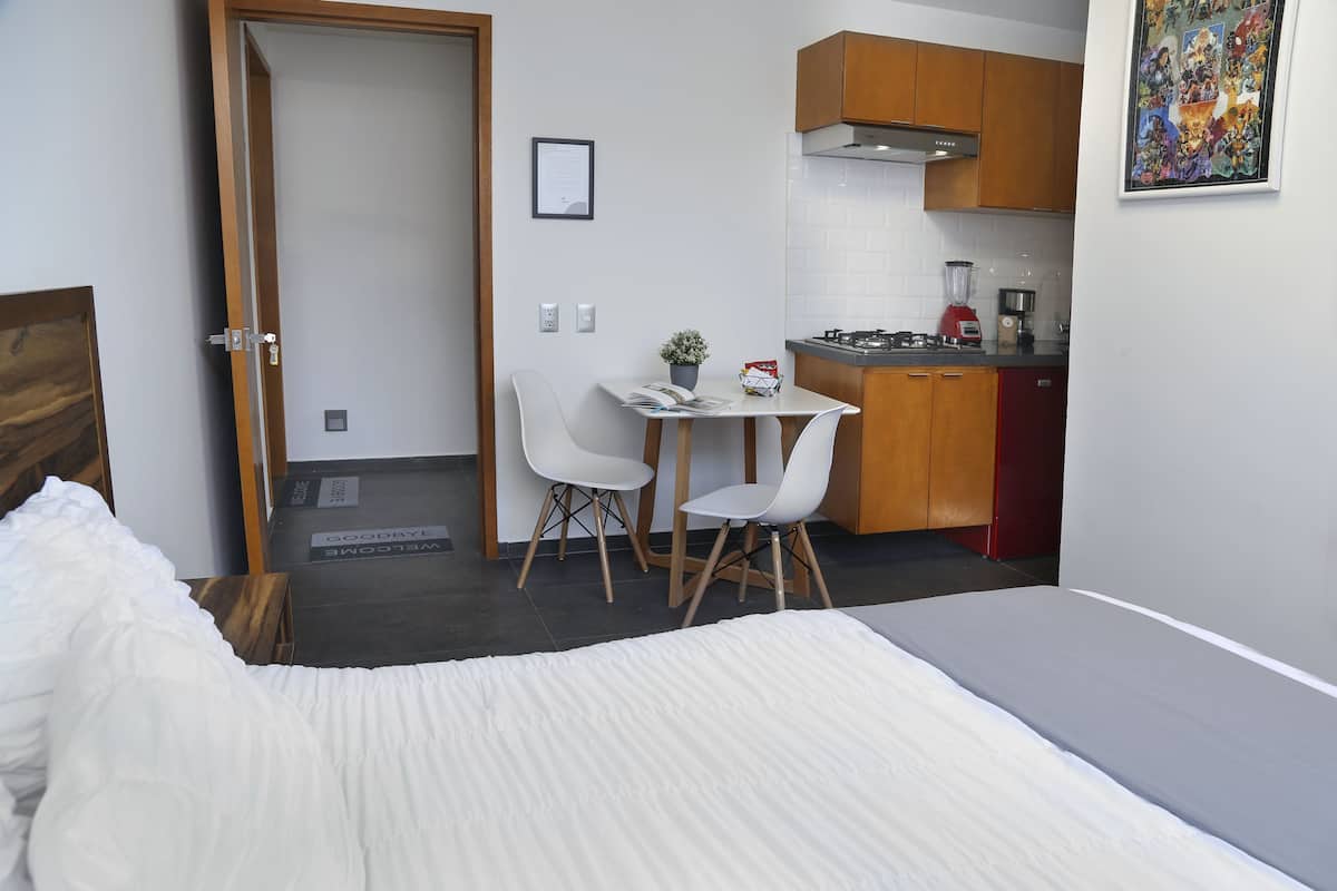 A compact hotel room with a bed, a small dining table with two chairs, a kitchen area with wooden cabinets, and a framed picture on the wall.