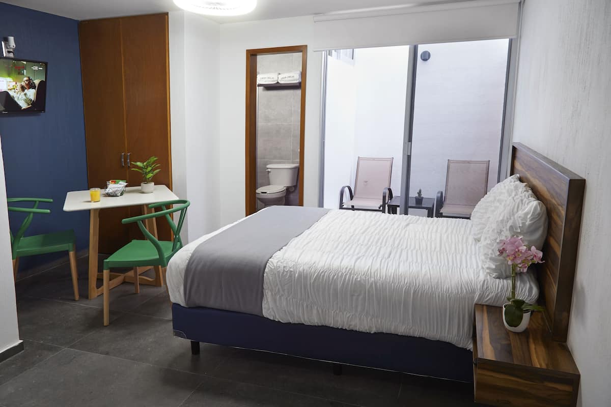 A bedroom with a bed, a desk, a chair, a small table, a potted plant, and a window.