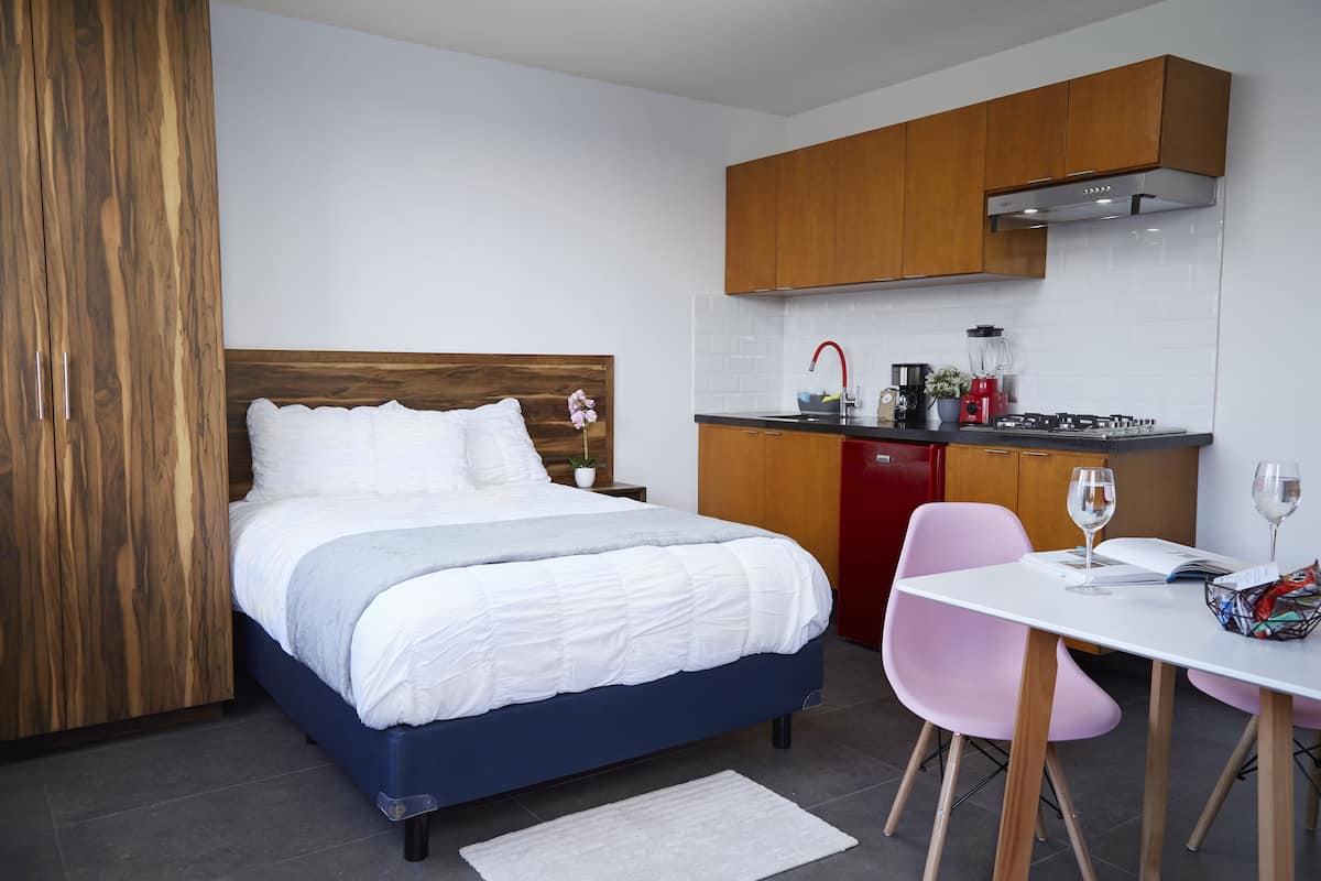 A bedroom with a bed, a small desk with a chair, a kitchen area with a sink, stove, and cabinets.