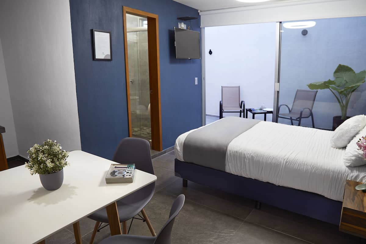 A hotel room with a bed, a desk with a chair, a potted plant, and a TV on the wall.