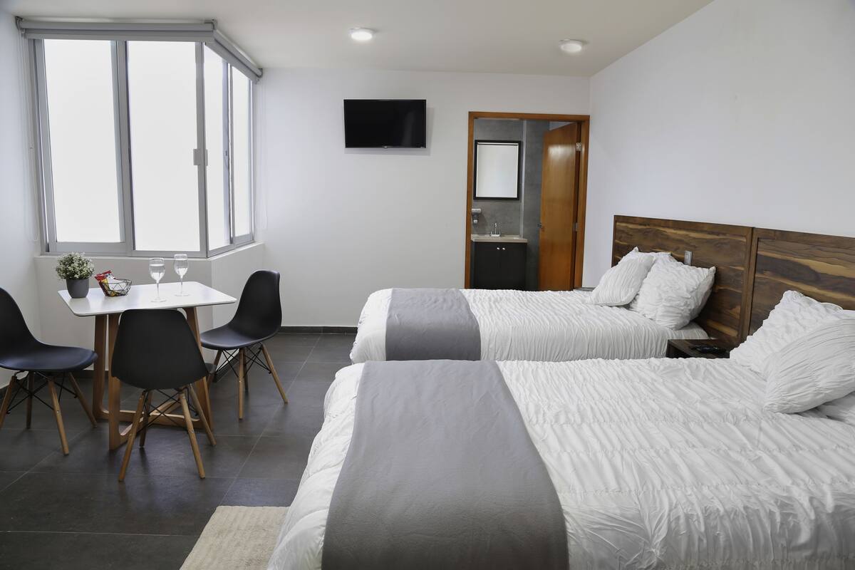 A hotel room with two beds, a desk with chairs, a TV, and a bathroom door.