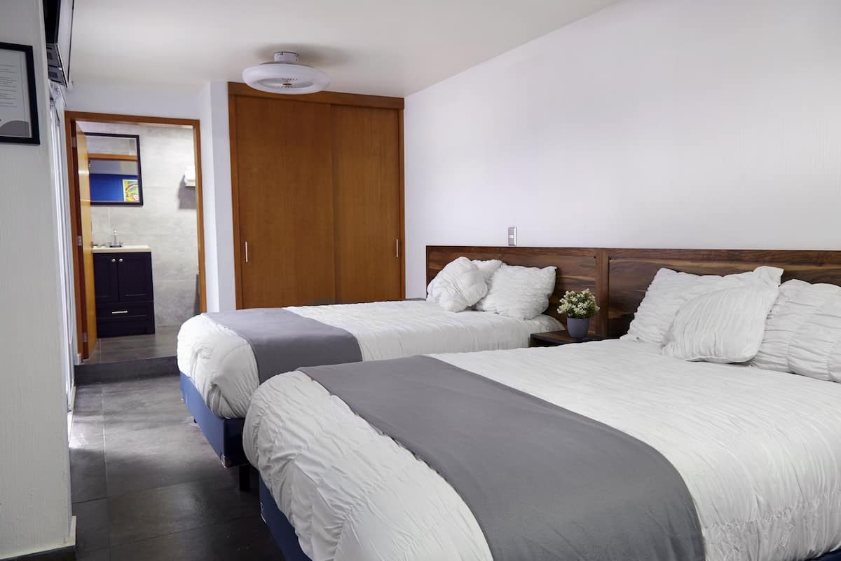 A hotel room with two beds, a wooden wardrobe, a small bedside table with a potted plant, and a bathroom visible through an open door.