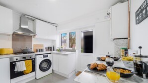 Una cocina moderna con estufa, horno y lavadora. Hay un espacio para desayunar con cruasanes, un tazón de cereal y un vaso de jugo de naranja.