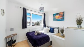 Un dormitorio pequeño con cama, mesita de noche, lámpara, ventana con cortinas, radiador, cuadro y planta en maceta.