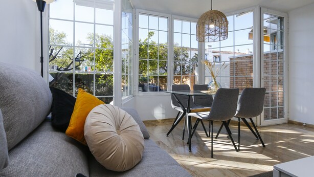 A modern living room with a sofa, a dining table with chairs, and large windows offering a view of the outdoors.