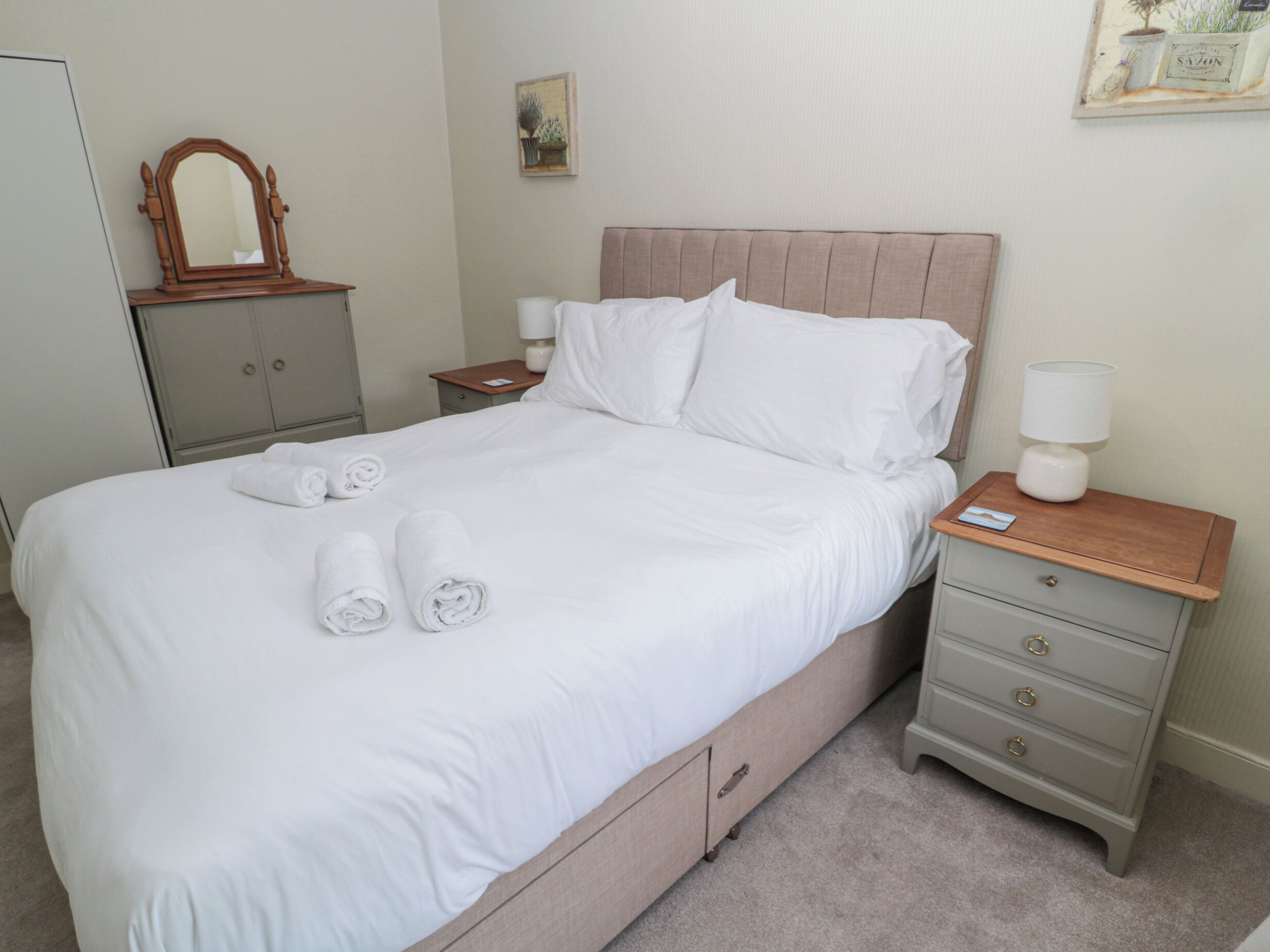 A neatly made bed with white linens and rolled towels, a wooden headboard, a bedside table with a lamp, and a small drawer unit.