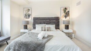 Une chambre moderne avec un grand lit, deux tables de chevet avec des lampes, un tapis moelleux et des œuvres d’art abstraites accrochées au mur.