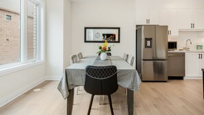 Un coin repas avec une table dressée pour six personnes, une cuisine moderne avec des meubles blancs, un réfrigérateur en acier inoxydable et une fenêtre donnant sur un bâtiment en briques.