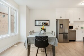 Un comedor con una mesa dispuesta para seis personas, una cocina moderna con armarios blancos, un refrigerador de acero inoxidable y una ventana con vista a un edificio de ladrillo.