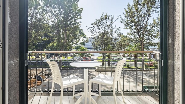 A balcony with two white plastic chairs and a small round table, overlooking a park with trees and a distant building. - King David Suites 201 by Homie (Funchal)