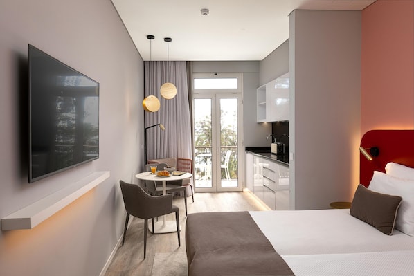 Ett modernt hotellrum med en platt-TV, en matplats med bord och stolar, en säng med sänggavel och ett köksutrymme.