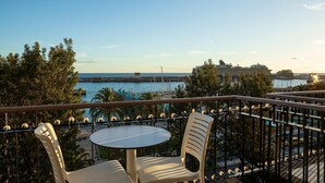 Ein Balkon mit zwei Stühlen und einem Tisch, der auf einen Yachthafen mit Booten und einem Kreuzfahrtschiff blickt.