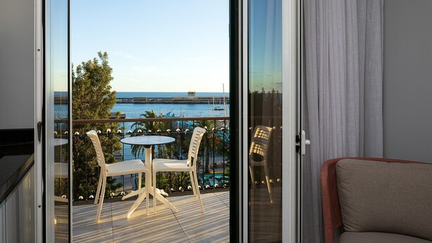 Ein Balkon mit Tisch und Stühlen, Blick auf das Meer, und ein Gebäude in der Ferne.