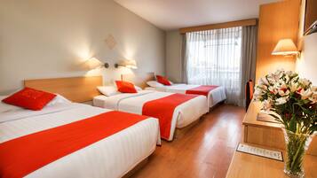 Habitación de hotel con tres camas, cada una con ropa de cama roja y blanca, un mostrador de recepción de madera y un jarrón con flores.