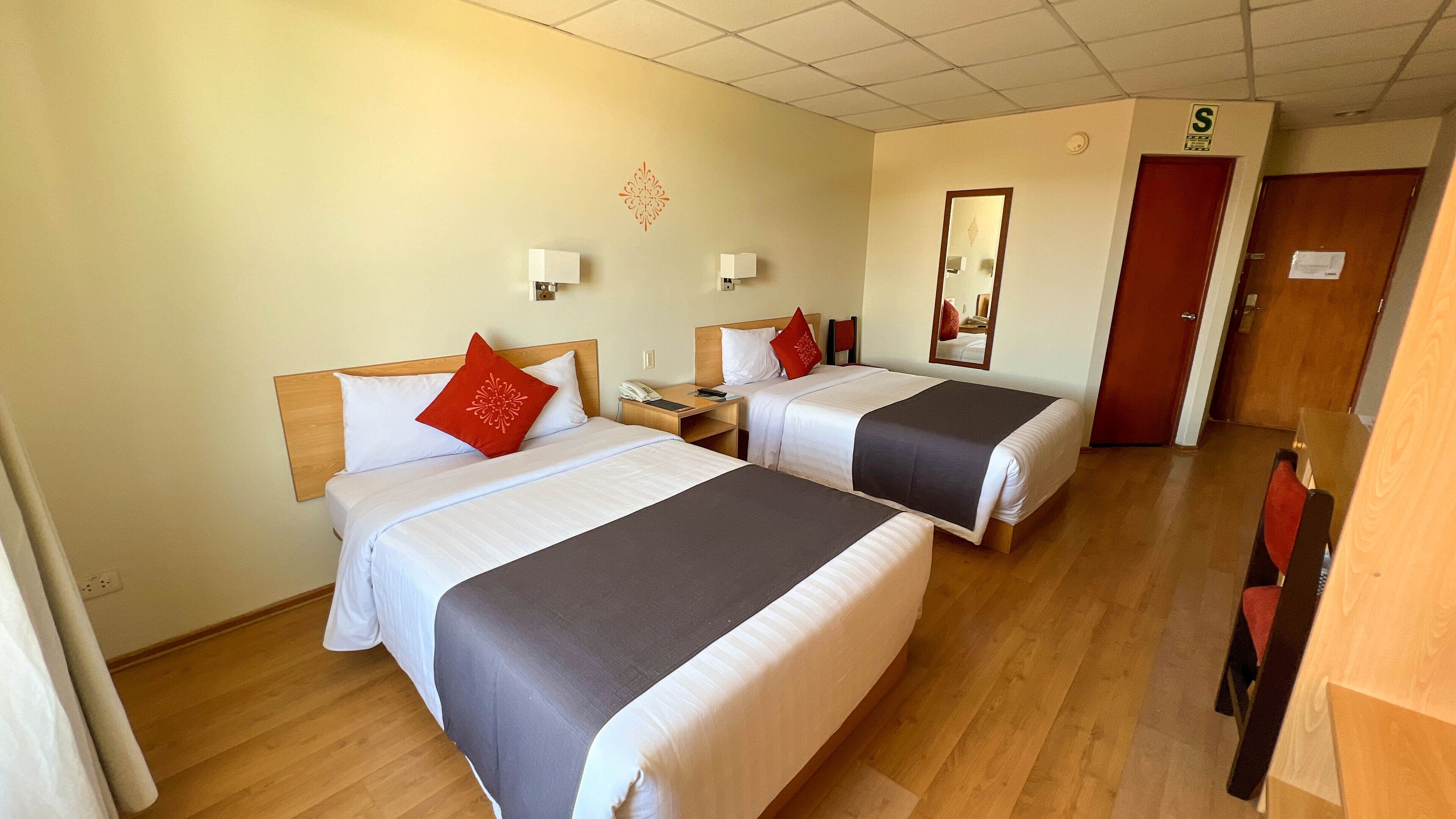A hotel room with two beds, each with a white and red pillow, a nightstand with a phone, and a mirror on the wall.