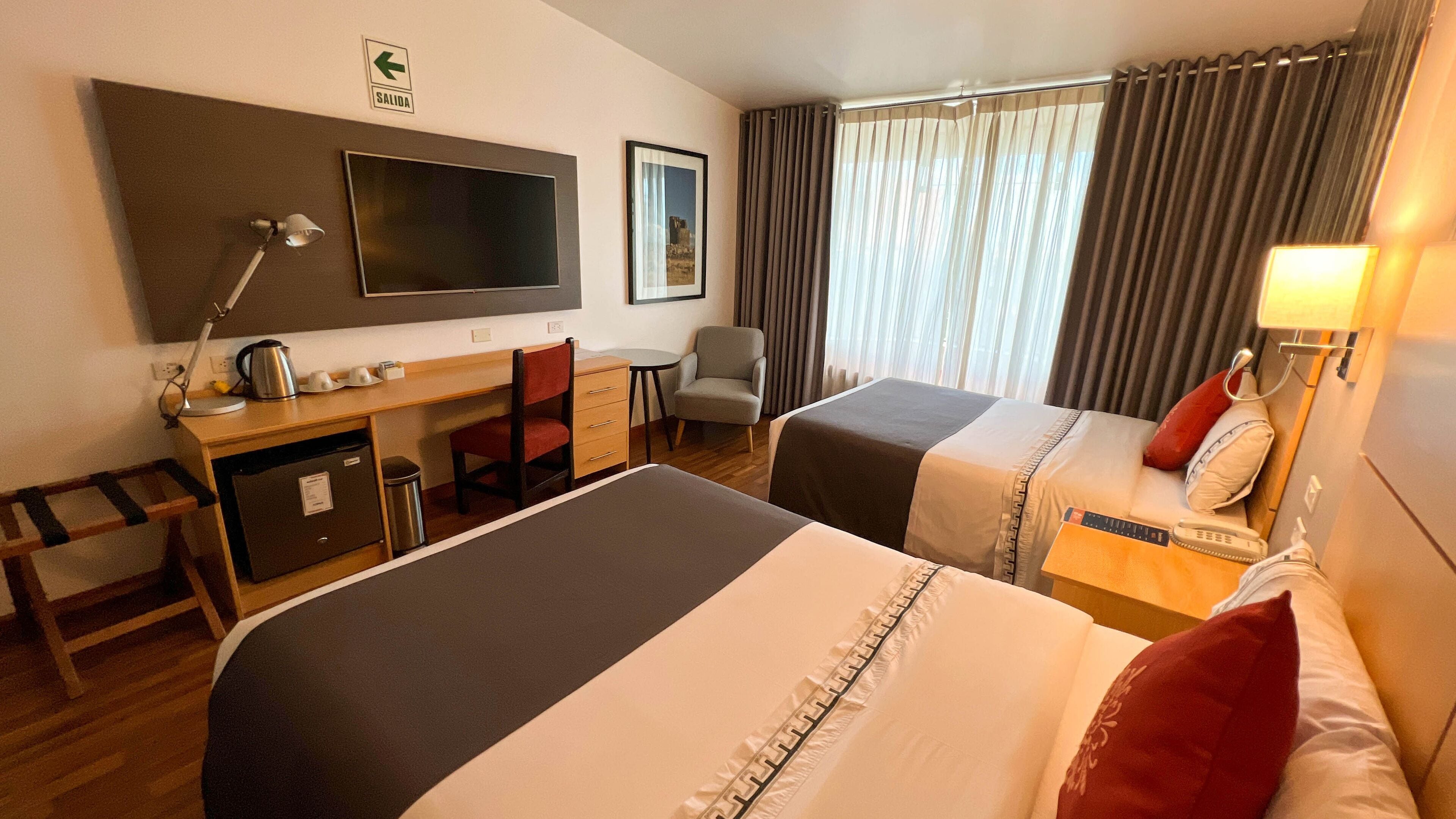 Une chambre d’hôtel avec deux lits, un bureau, une chaise, une télévision et une fenêtre avec des rideaux.