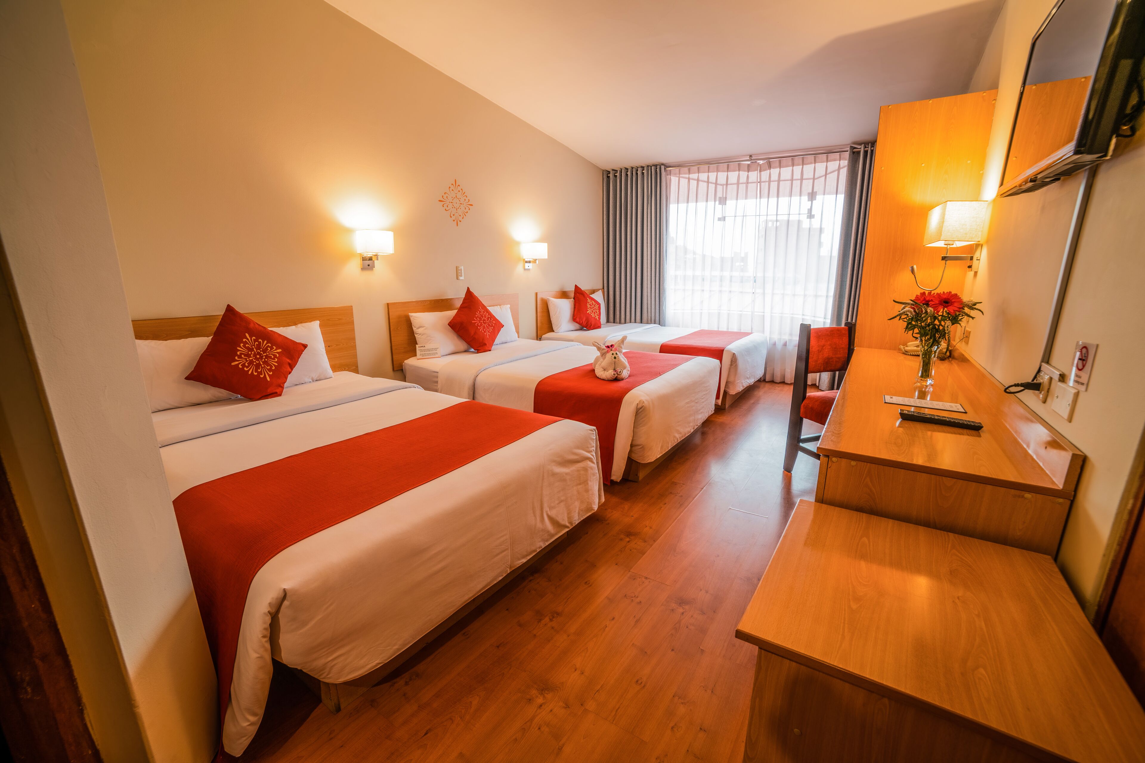 Habitación de hotel con tres camas, cada una con ropa de cama roja y blanca, un escritorio de madera y un espejo.