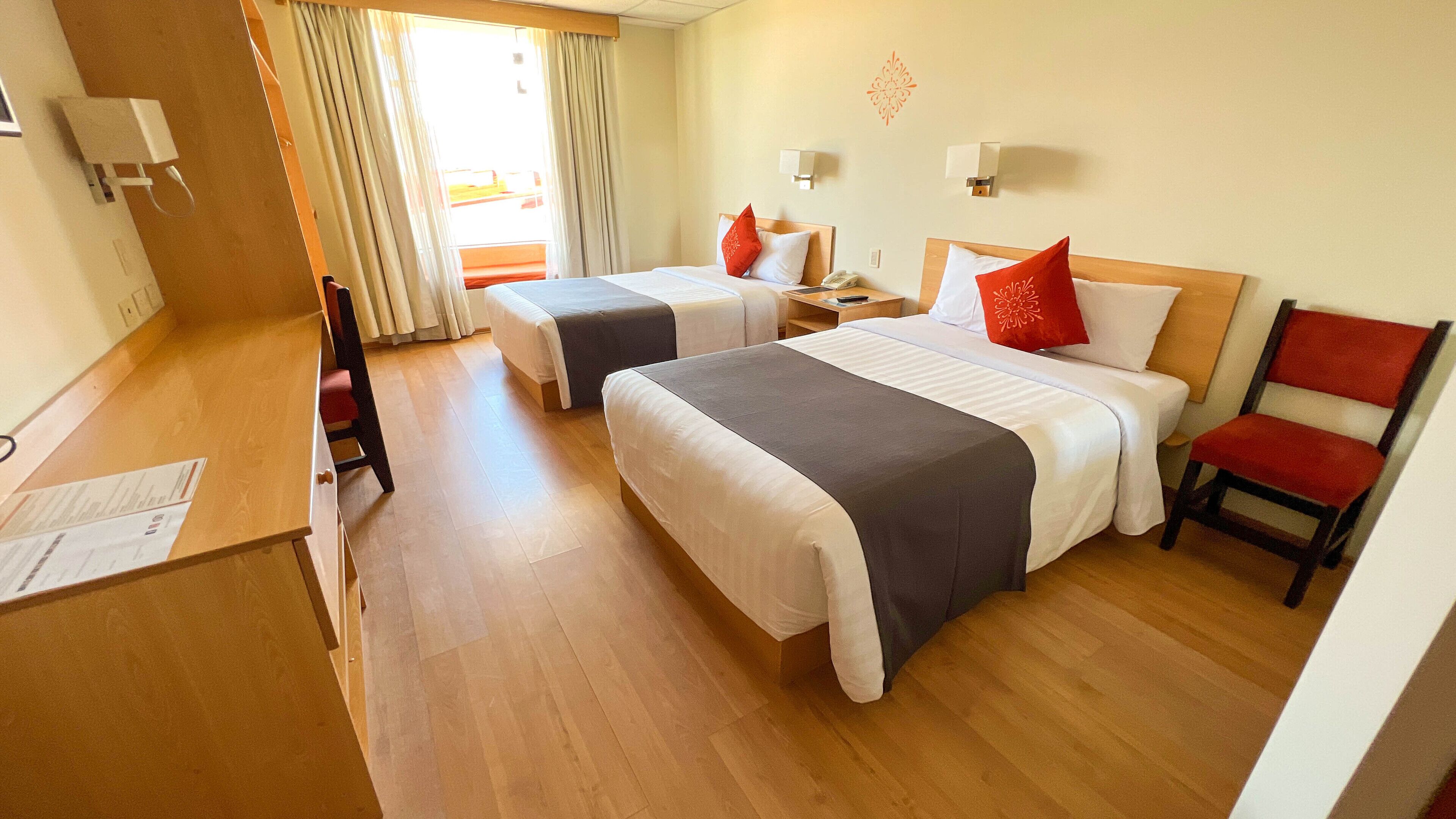 Habitación de hotel con dos camas, un escritorio, una silla y una ventana con cortinas.