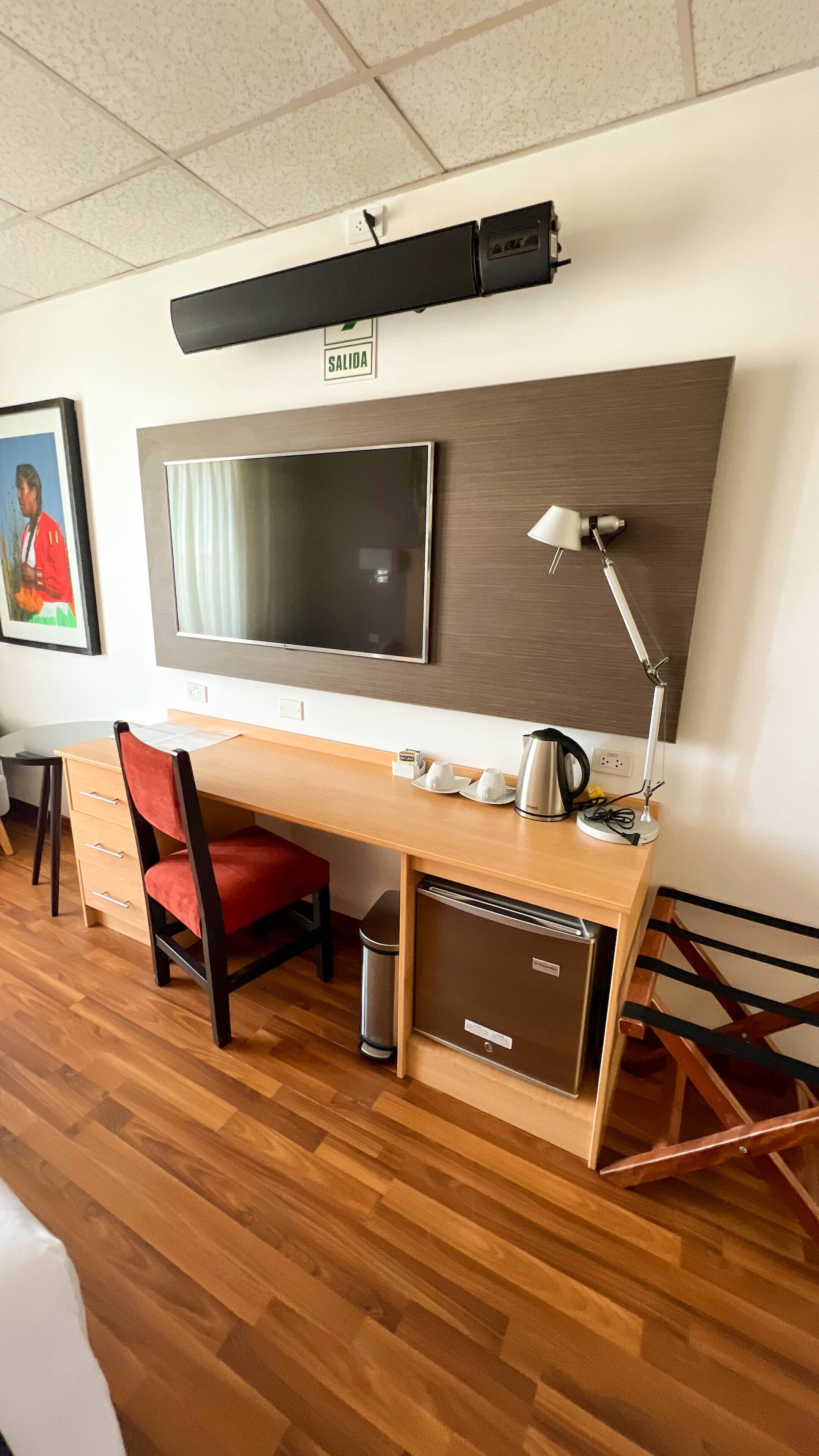 Habitación de hotel con un escritorio de madera, una silla, un televisor montado en la pared, una lámpara, un cesto de basura y un cartel que indica la salida.