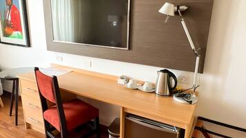 Habitación de hotel con un escritorio de madera, una silla, un televisor montado en la pared, una lámpara, un cesto de basura y un cartel que indica la salida.
