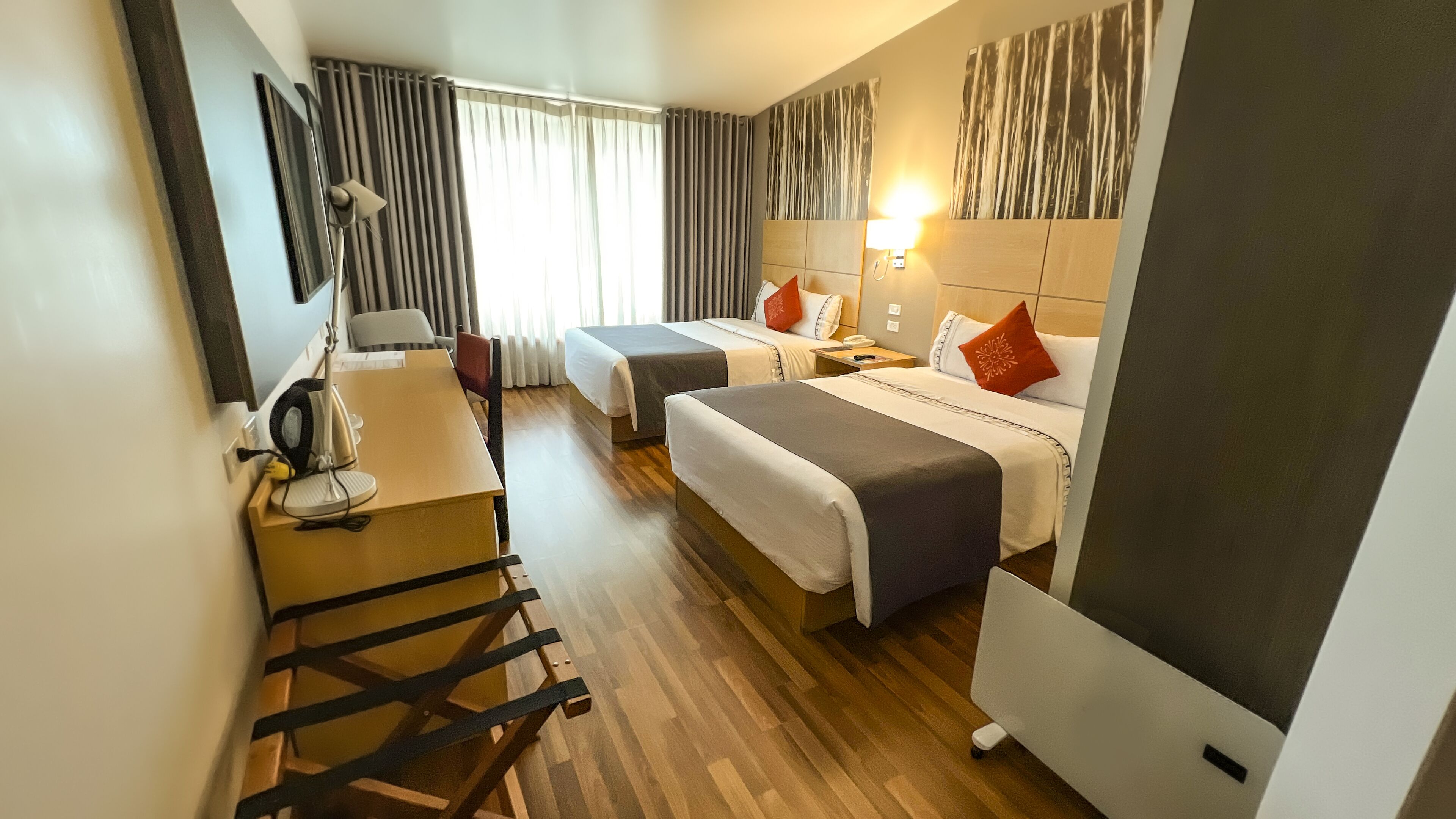 Habitación de hotel con dos camas, un escritorio con silla, una lámpara y un ventanal grande con cortinas.