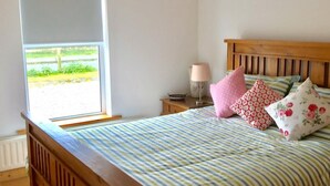 A bedroom with a wooden bed, a window with a view of greenery, a bedside table with a lamp, and a nightstand. - 2 min's to beautiful beach, amazing home for 8 peeps huge garden. Suits families (Portsalon)