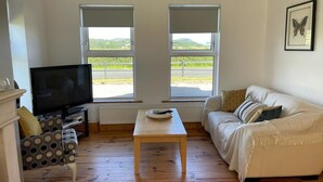 A living room with a TV, a sofa, a coffee table, and two windows with a view of greenery. - 2 min's to beautiful beach, amazing home for 8 peeps huge garden. Suits families (Portsalon)