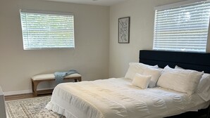 Un dormitorio con cama, ventilador de techo, dos ventanas con persianas, una mesita de noche y una alfombra.