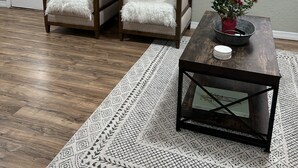 Un salón con dos sillones, una mesa de centro, un tapete estampado, una puerta con panel de vidrio y un cuadro enmarcado en la pared.