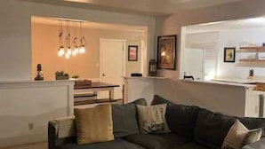 A living room with a dark-colored sofa, a small ottoman, and a patterned rug. There is a kitchen area visible in the background with a white door and a hanging light fixture. - Three Kings Ranch (Indianapolis)