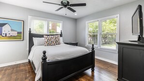 A bedroom with a bed, a ceiling fan, two windows, a painting, and a TV on a cabinet. - Urban Cowboy  (Fort Worth)