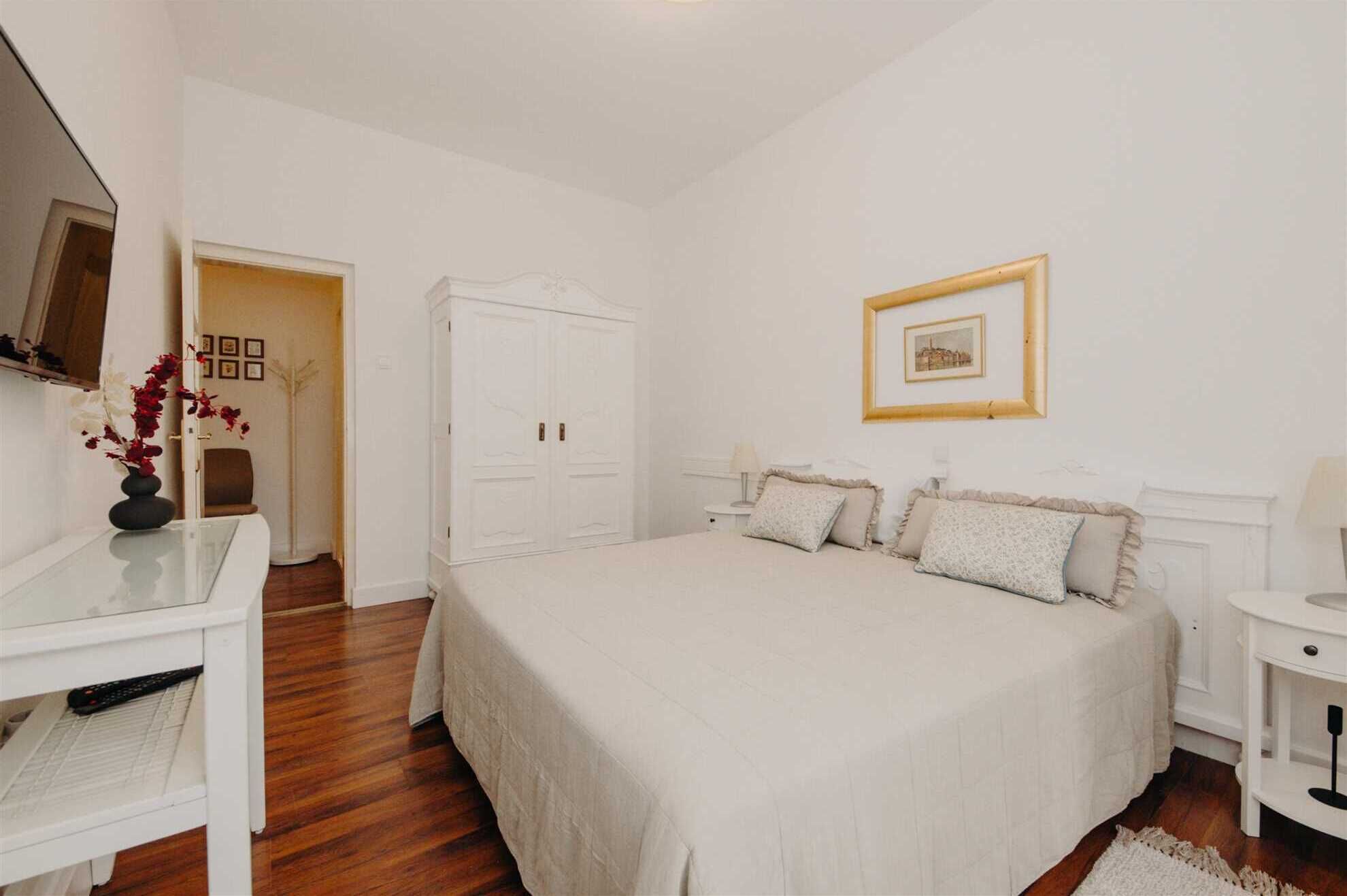 Een slaapkamer met een groot bed, een bureau en een stoel. Er is een garderobe en een lijst met een afbeelding aan de muur.