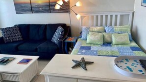 A bedroom with a bed, a nightstand, a dresser, a sofa, and a wall with a sunset painting. - Ocean Front Studio on Boardwalk (Virginia Beach)