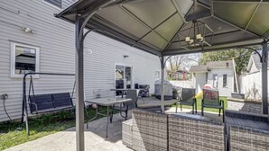 En overdækket patio med et bord og stole, en gyngestativ og et overdækket siddeområde med udsigt til et hus i baggrunden.