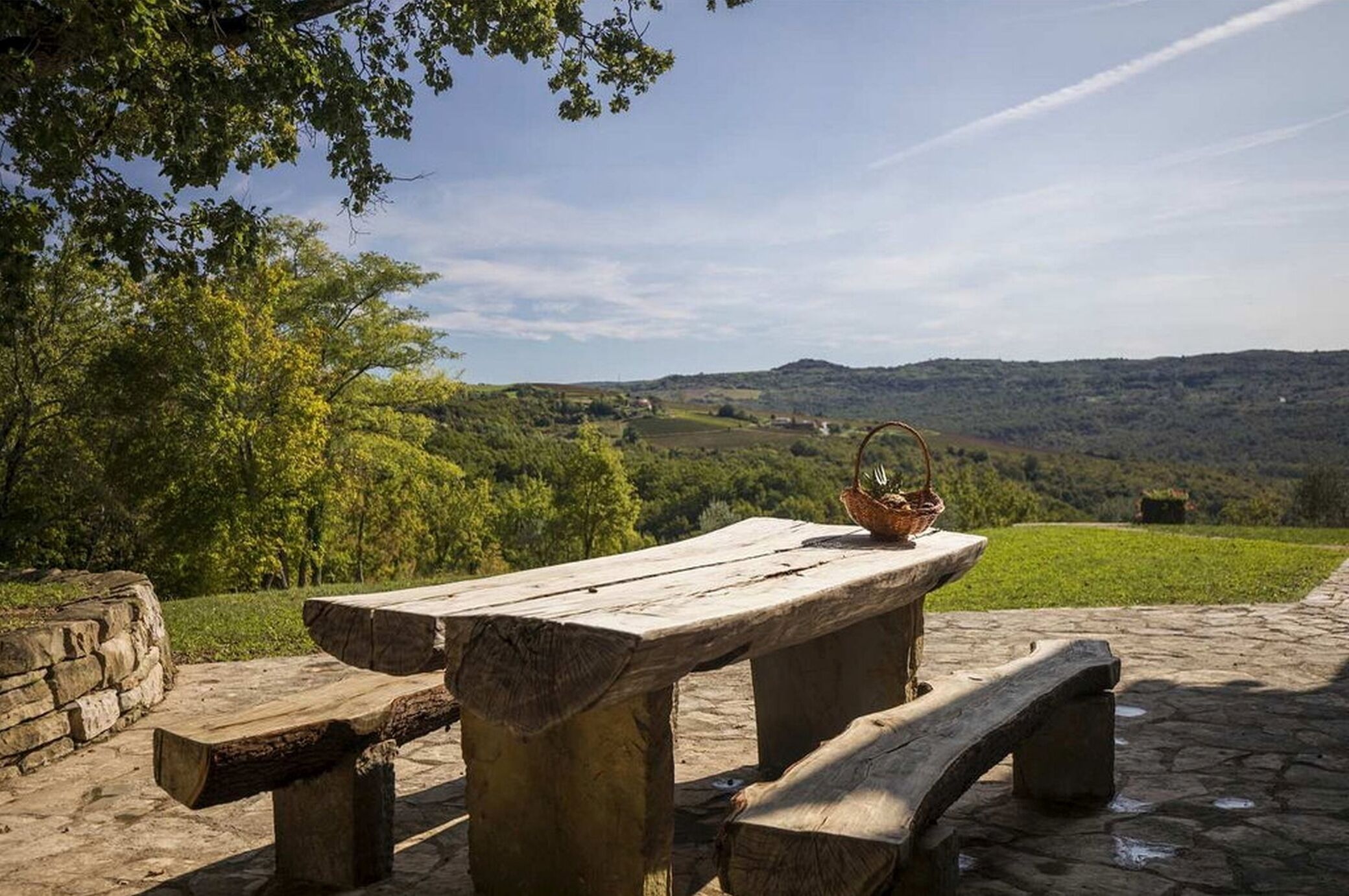 A rustic picnic table with a basket on it, set on a stone patio with a view of rolling hills and trees in the background.