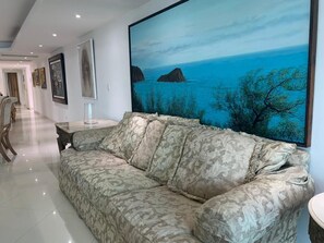 A living room with a patterned sofa, a coffee table, and a large painting of a coastal landscape.