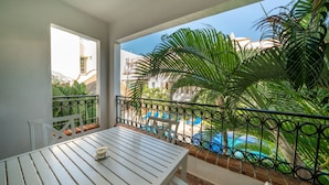 A balcony with a white table and chairs, a view of a swimming pool, and surrounding greenery. - Condo Gaviota - Tropical Oasis (Playa del Carmen)