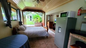 A room with a bed, a small table, a refrigerator, and a window overlooking a garden.