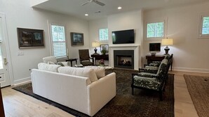 A spacious living room with a white sofa, patterned armchairs, a fireplace, and a flat-screen TV mounted above it.