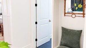 A room with a white door, a green cushioned bench, a wall-mounted mirror, and a potted plant.