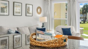 Un salon comprenant un canapé, des fauteuils, une table basse, des tableaux encadrés, une lampe et une fenêtre avec des rideaux.