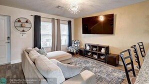 Een woonkamer met een zithoek, een aan de muur bevestigde televisie en een eethoek met stoelen.