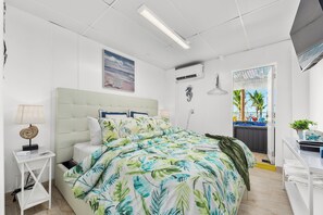 A bedroom with a bed, a nightstand, a lamp, a wall-mounted air conditioner, and a window with a view of palm trees. - The Resort at Bathtub Beach: 19BR/11BA Ocean2River, Pool, Elevator, Dock, Beach! (Stuart)