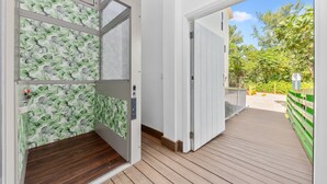 Une terrasse en bois menant à une porte blanche ornée d’un motif de feuilles vertes, un ciel dégagé et une clôture verte.
