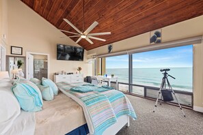 A bedroom with a large bed, a ceiling fan, a TV, and a view of the ocean.