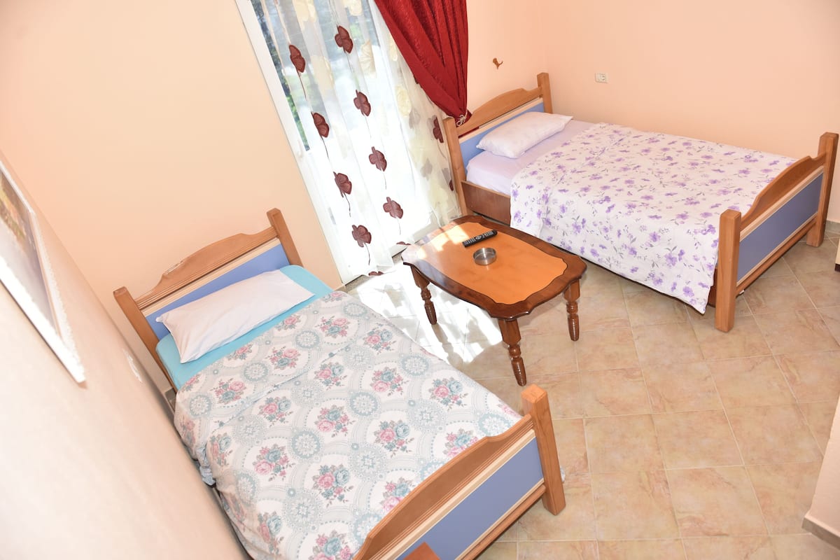 Two wooden beds with floral bedding, a small wooden table with a remote control, and a red curtain.