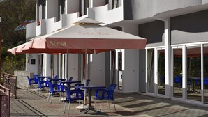 Een gebouw met een terras, buitenmeubilair met tafels en stoelen, en een grote parasol.