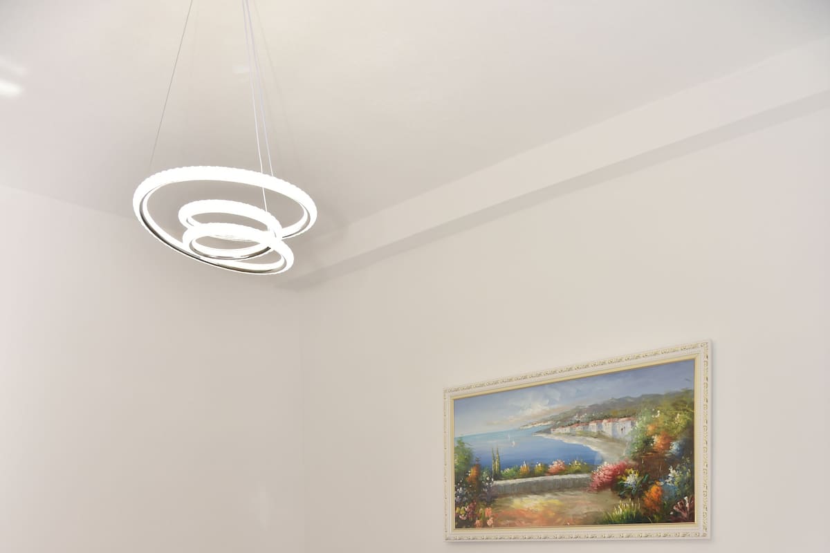 A bedroom with a bed, a grey sofa, a nightstand, a painting, and a modern light fixture.
