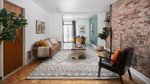 A modern living room with a brick wall, a wooden floor, a brown sofa, a coffee table, and a dining area in the background.